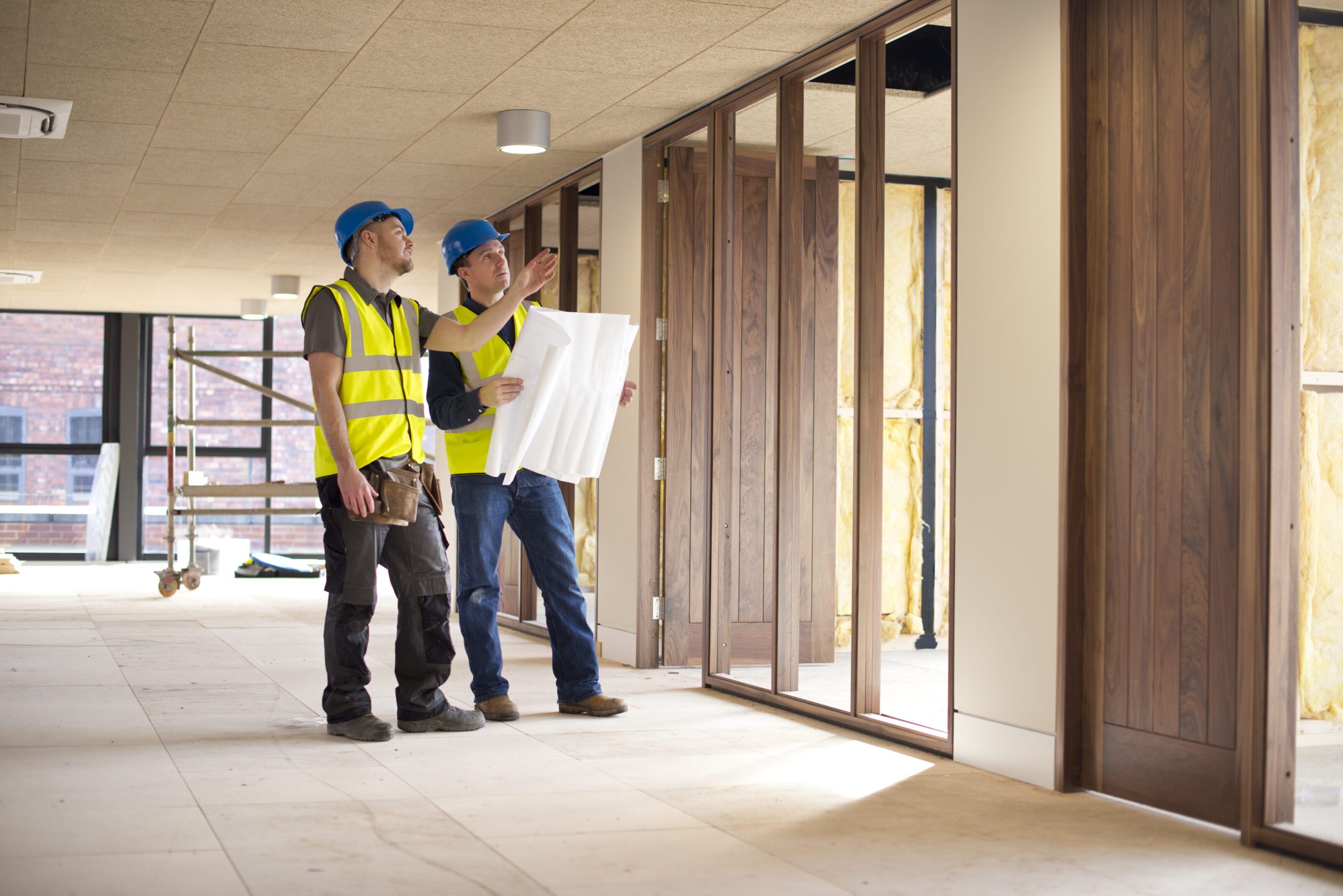 Two construction workers reviewing building plans inside a framed structure.