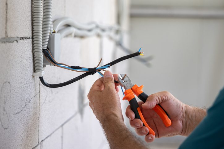 Hands cutting electrical wires with wire cutters near a wall.